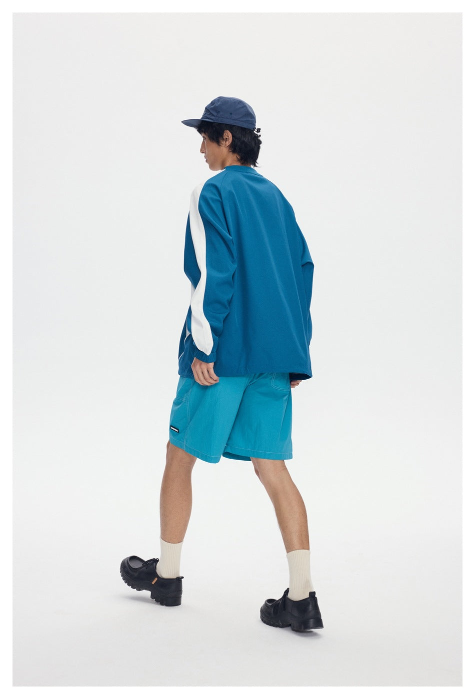 Back view of model wearing blue pullover with white sleeve stripe, turquoise shorts, navy cap and black shoes
