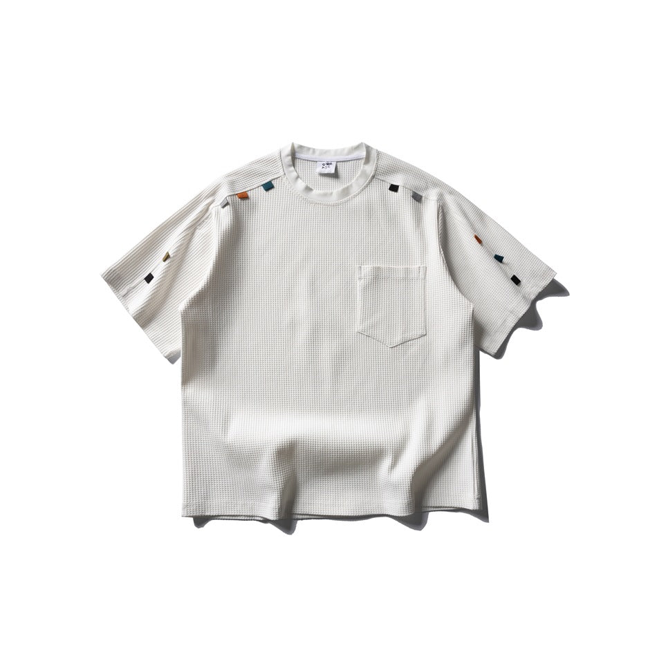 Front view of an ivory short-sleeve waffle-textured T-shirt with a chest pocket and small colorful fabric tabs.