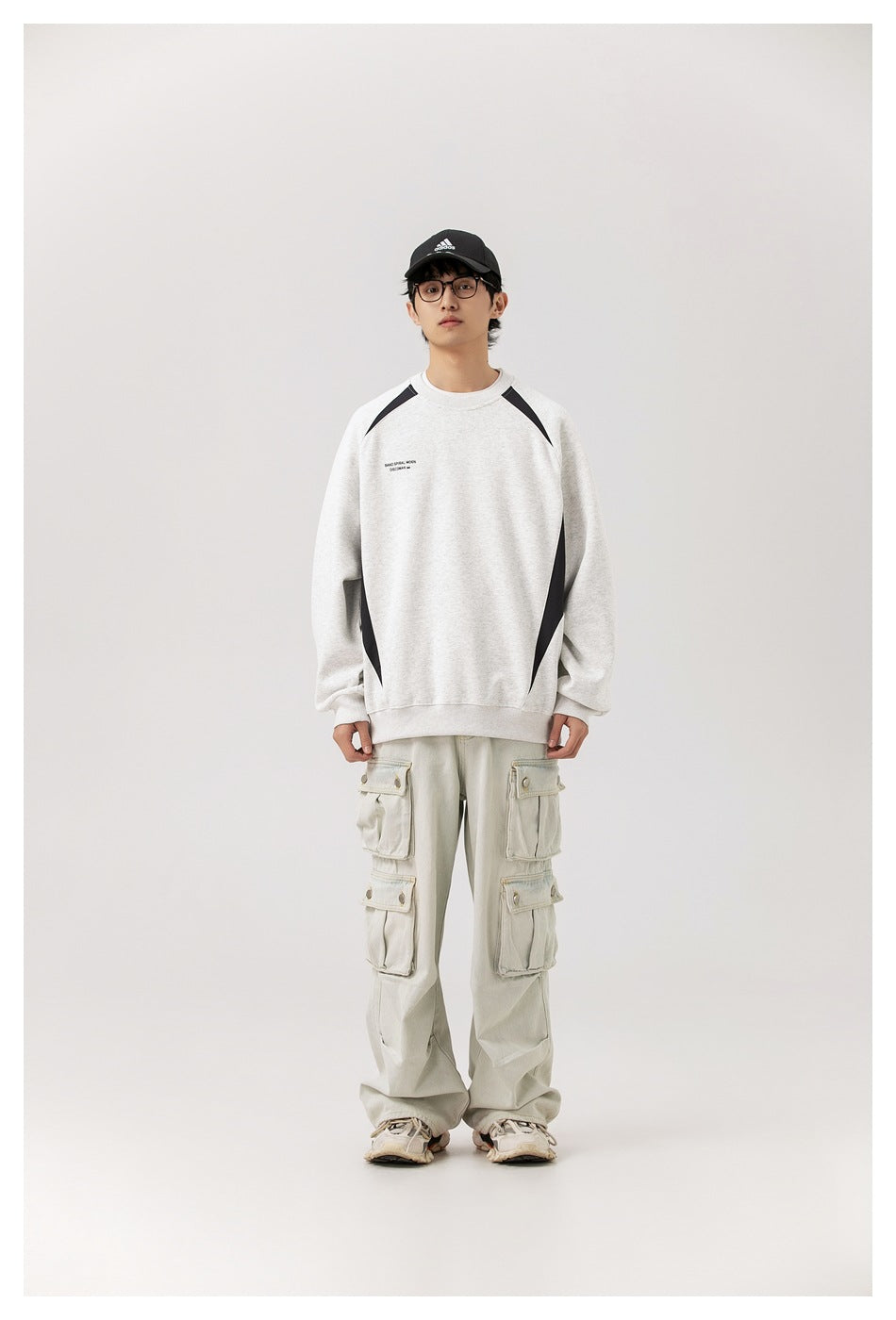 Model wearing gray crew-neck sweatshirt with black contrast panels, front view, with light cargo pants and cap