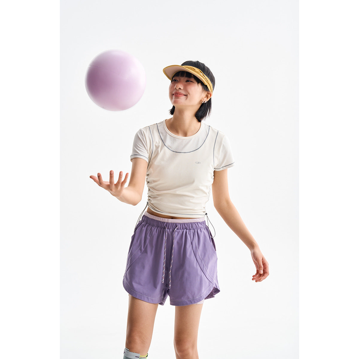Model wearing a cream cropped athletic tee with gray piping and loose lavender drawstring shorts.