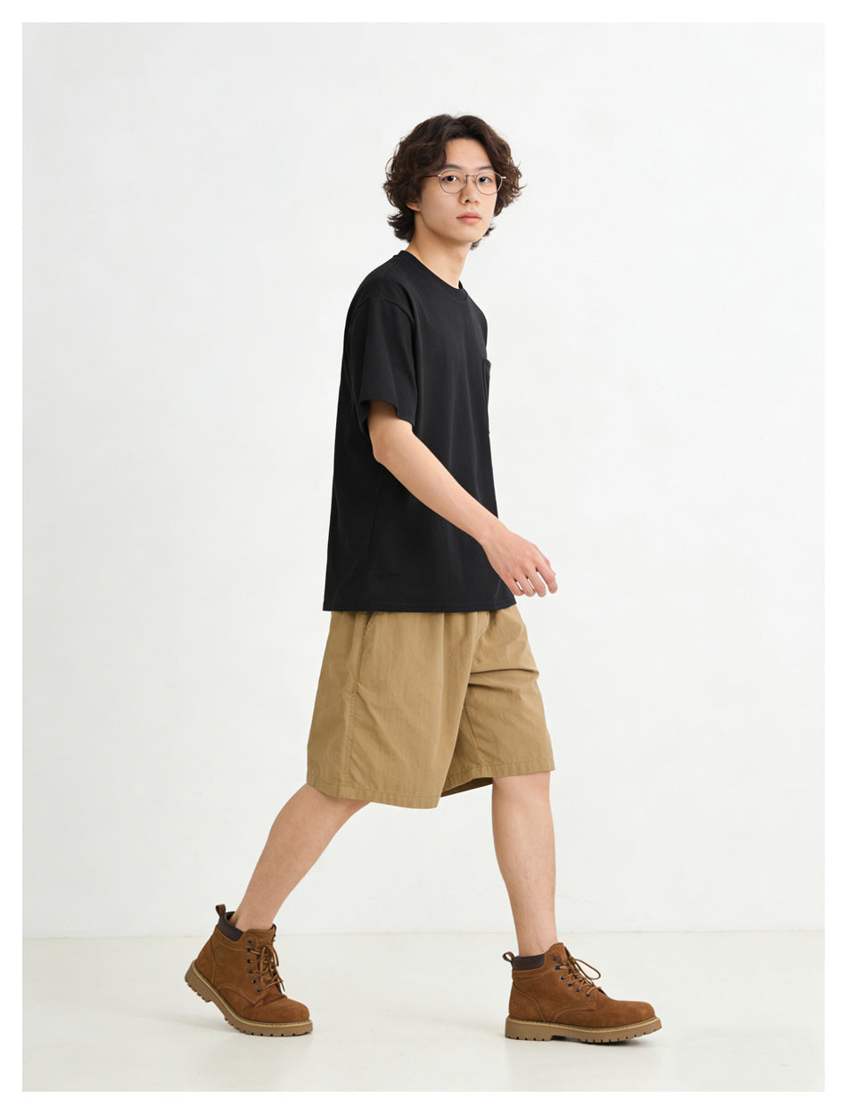 Model walking in a black short-sleeve crew-neck T-shirt with a chest pocket and tan shorts.