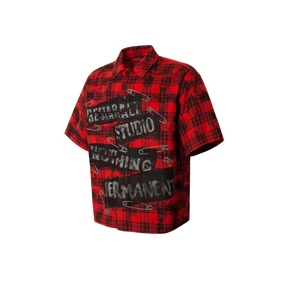 Angled front view of a red-and-black plaid short-sleeve shirt with English lettering and safety-pin graphics.
