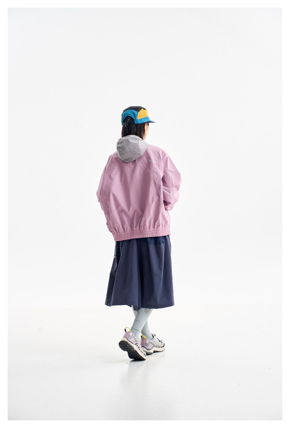 Rear view of a model in a pink jacket with a gray hood, navy skirt, and color-block cap.