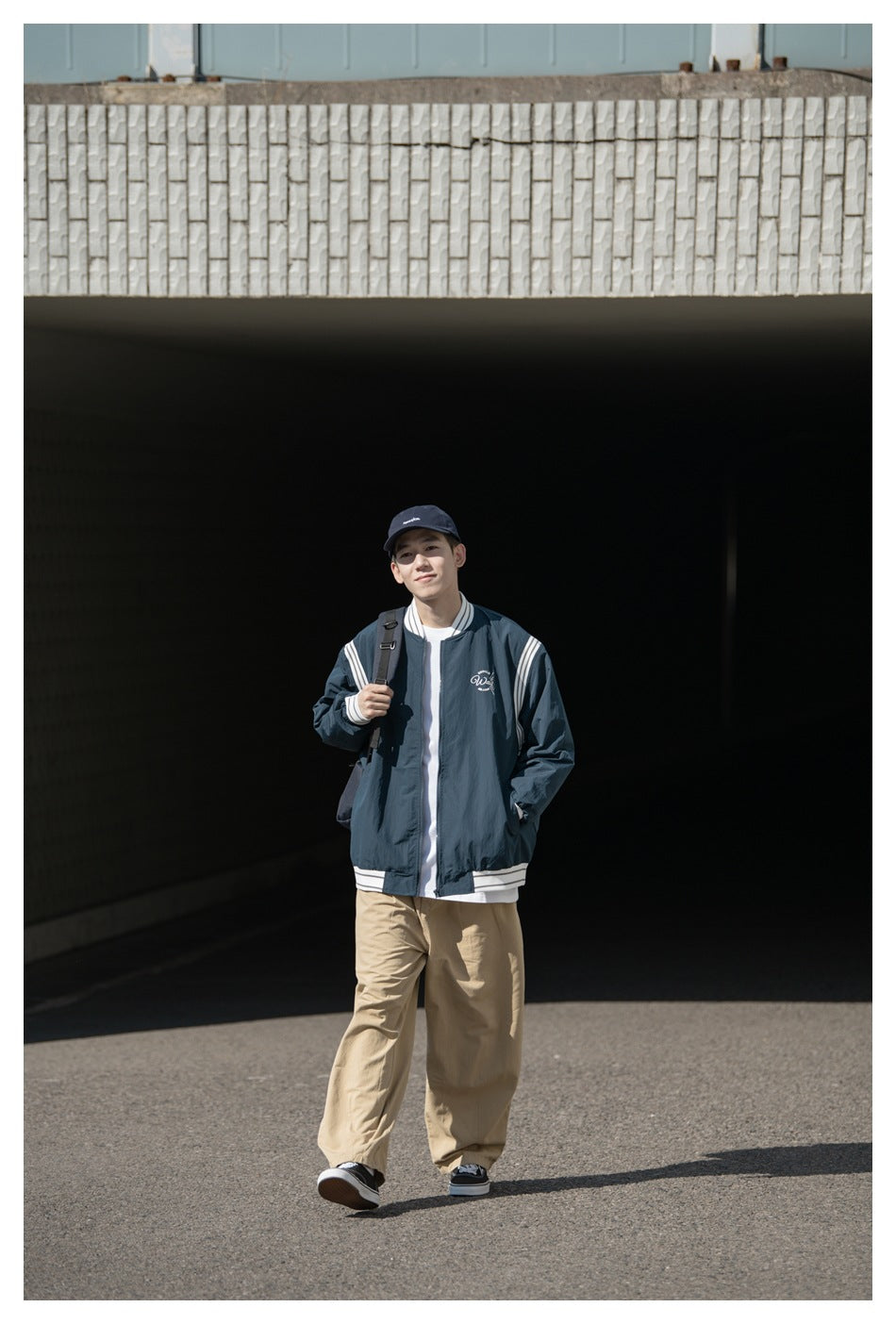 Smiling man wearing a navy bomber jacket with white stripes, khaki wide pants, cap and backpack in a tunnel