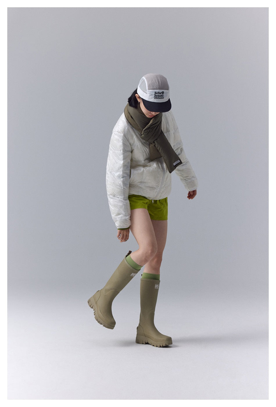 Model wearing a white quilted jacket, olive scarf, lime-green shorts, and tall olive boots, shown from the front.