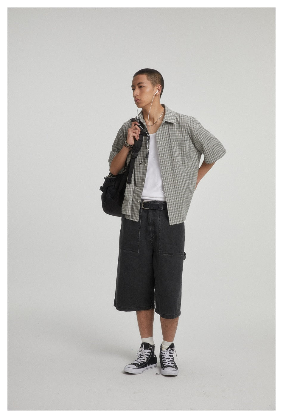 Model wearing a gray plaid short-sleeve button-up shirt over a white tank and black knee-length shorts, front three-quarter