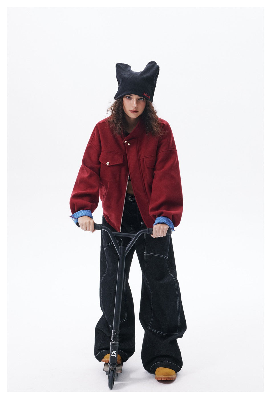 Front view of a model standing with a scooter in a red jacket, blue checked cuffs, black wide-leg jeans and a black beanie.