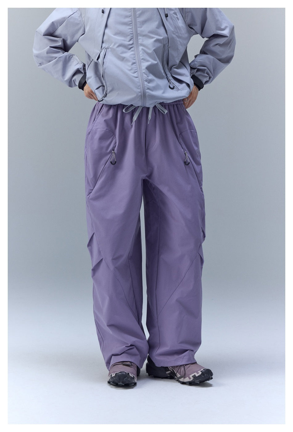 Front view of loose lavender-purple pants with an elastic drawstring waist and zip pockets, worn with a light gray jacket.