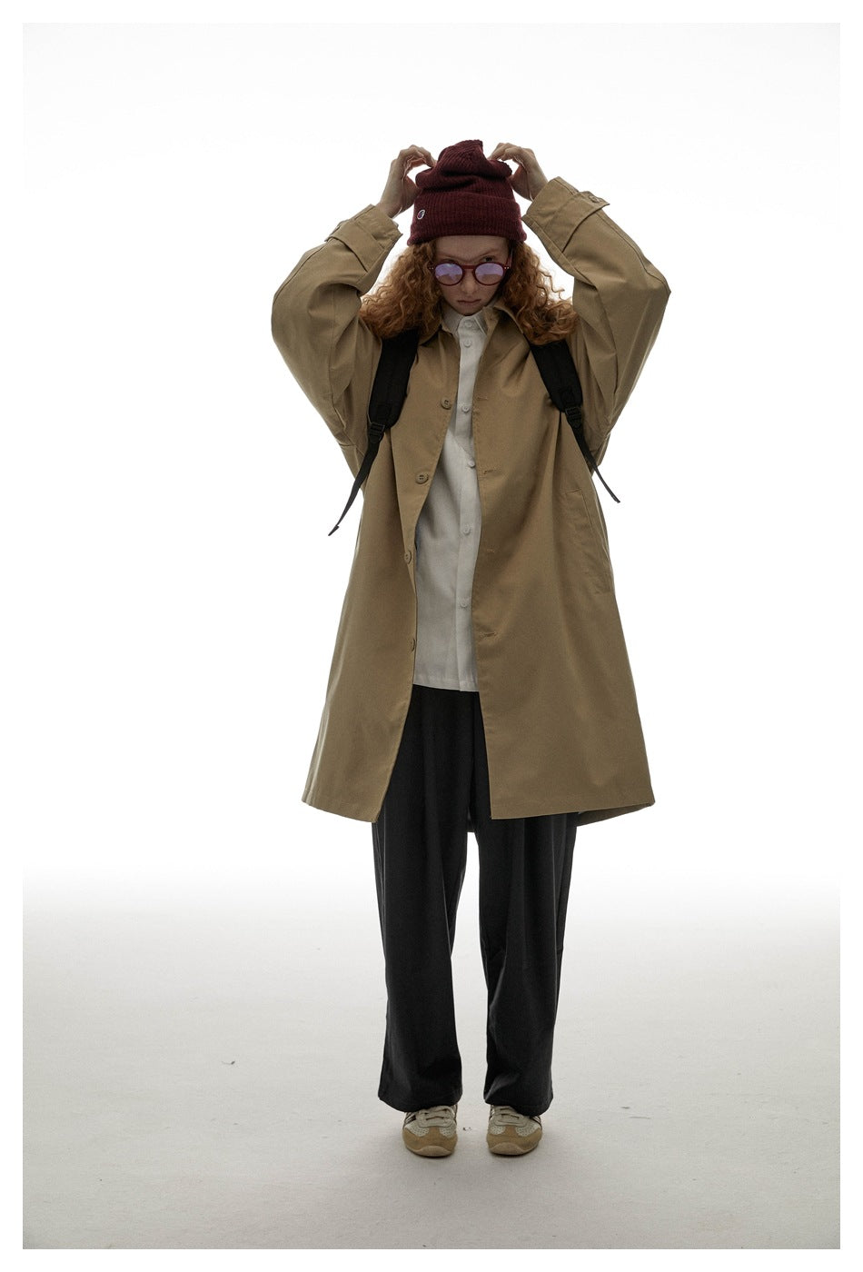 Model wearing an oversized tan button-front coat over a white shirt and black trousers, adjusting a burgundy beanie.