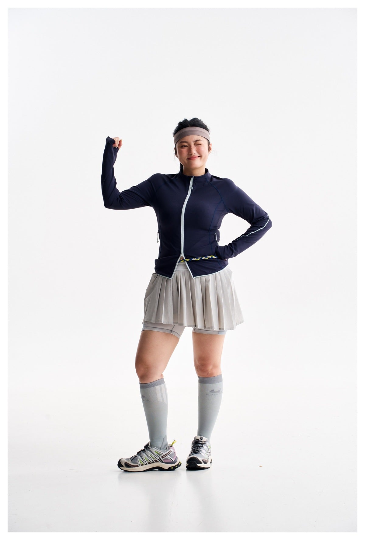 Model posing in a navy zip-up athletic jacket, gray pleated skort, and gray knee-high socks.