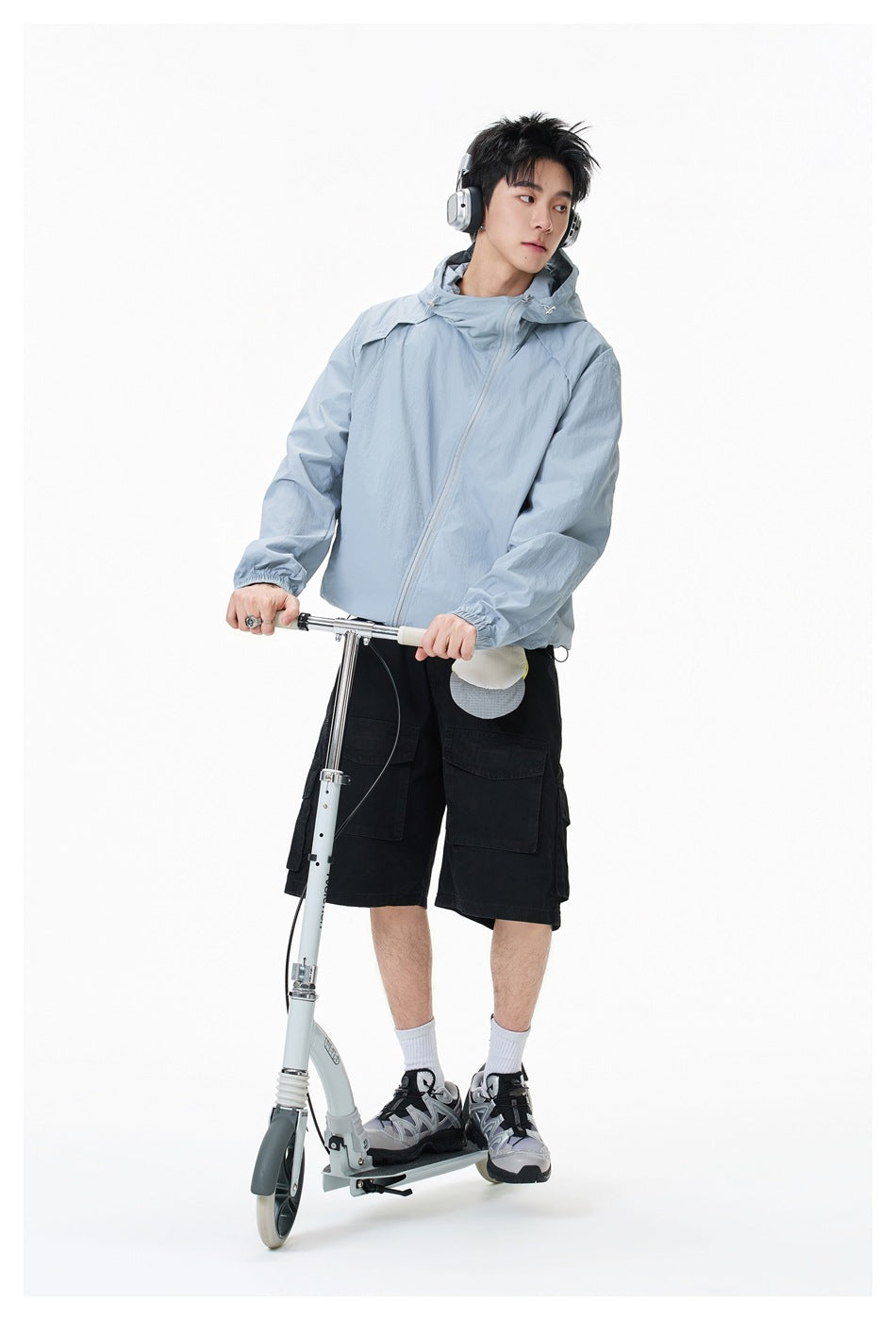 Model wearing a pale blue hooded jacket and black cargo shorts while standing on a scooter.