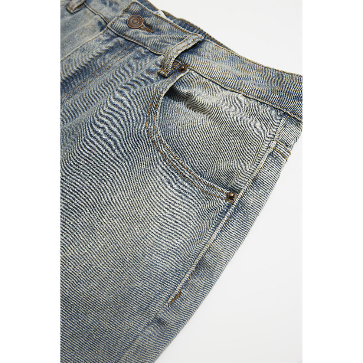 Close-up of light blue jeans showing a curved front pocket, waistband button, belt loop and contrast stitching.