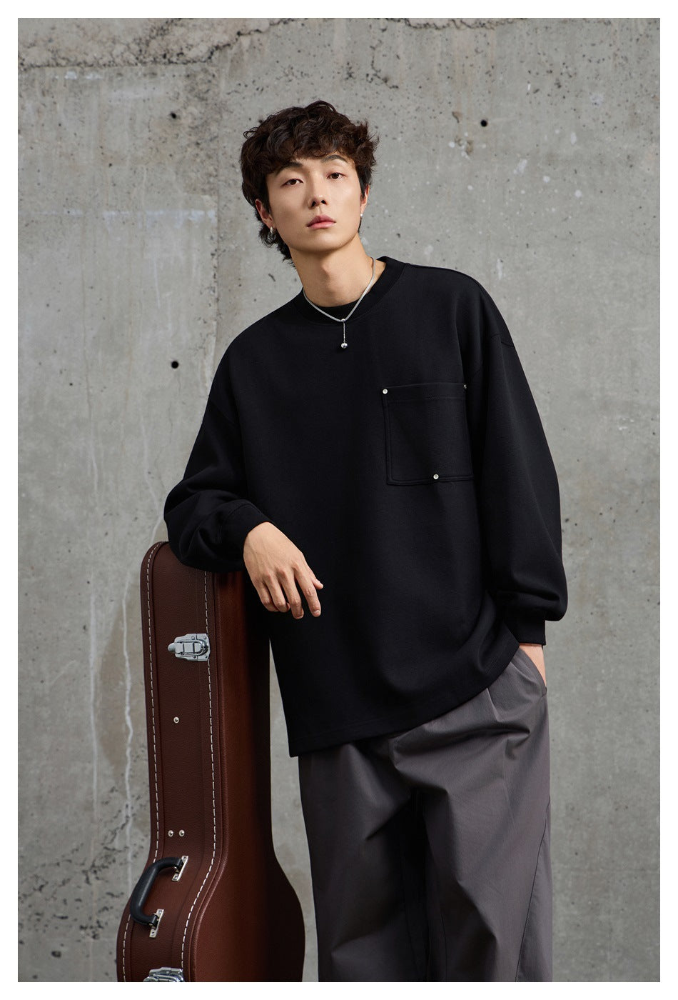 Front view of a model in a black oversized crewneck sweatshirt with a square chest pocket and loose gray trousers.
