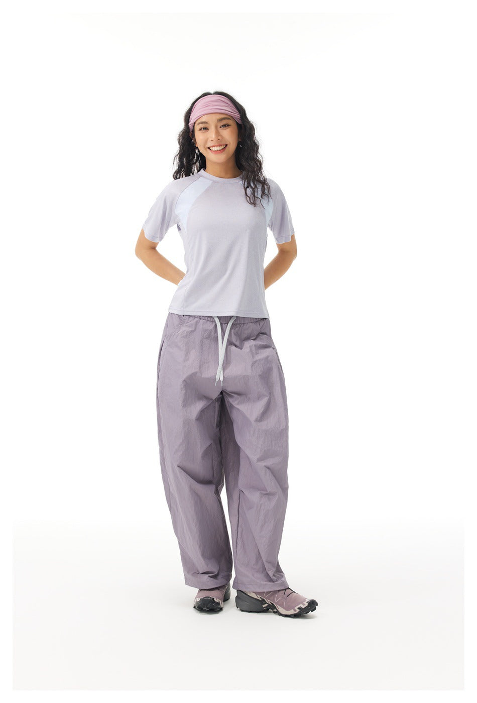 Model in a pale lavender color-block T-shirt and loose lavender-gray drawstring pants, standing with hands behind her.