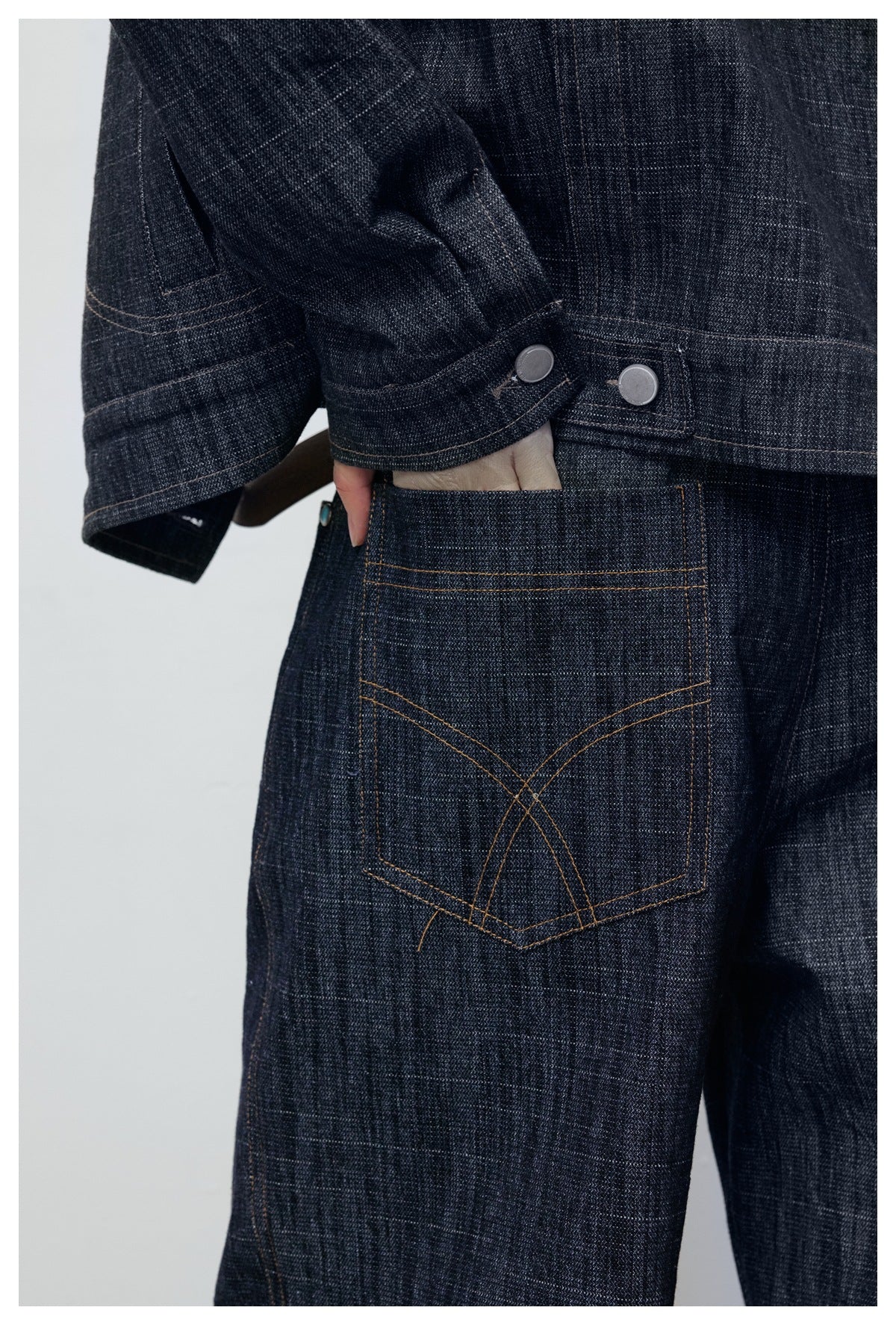 Close-up of a person wearing dark indigo pants with a stitched back pocket and a jacket cuff with snap tabs.