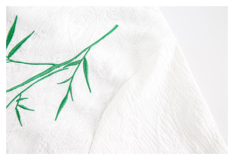 Close-up of green branch embroidery and raised white fabric texture beside a curved garment seam.