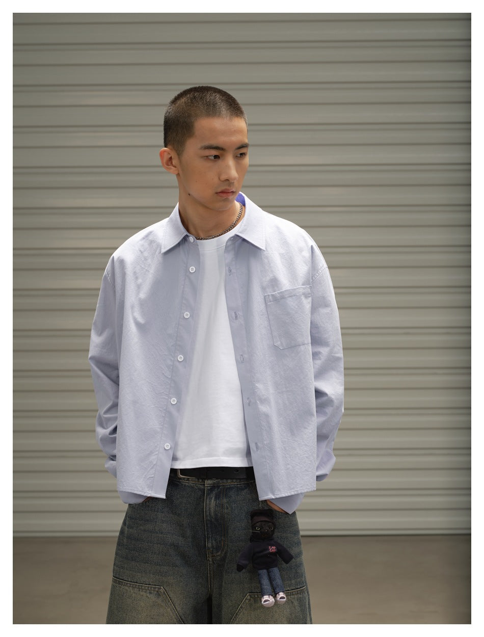 Waist-up three-quarter view of a model wearing a pale blue button-up shirt over a white tee, with a single chest pocket.