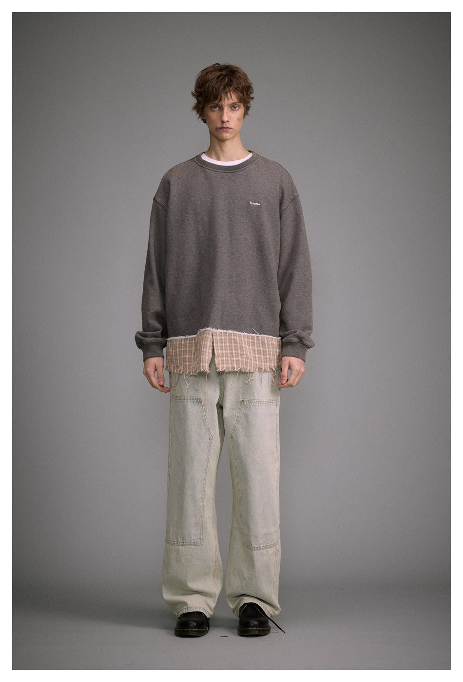 Full-length front view of a man wearing a charcoal oversized sweatshirt with a frayed plaid hem and loose pale jeans.