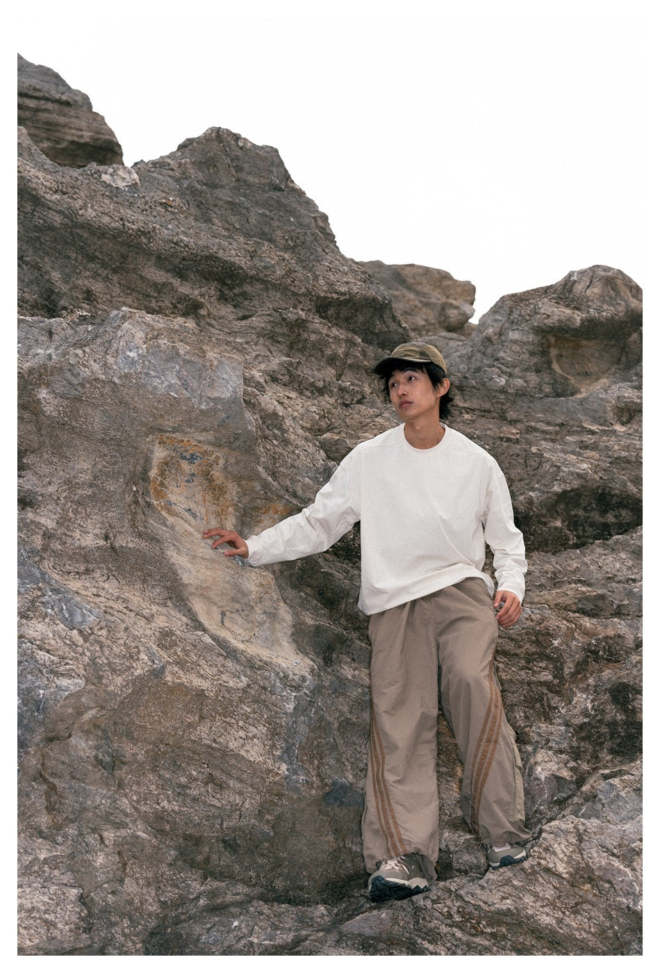 Model among rocks wearing an off-white long-sleeve top and taupe pants with brown side stripes.