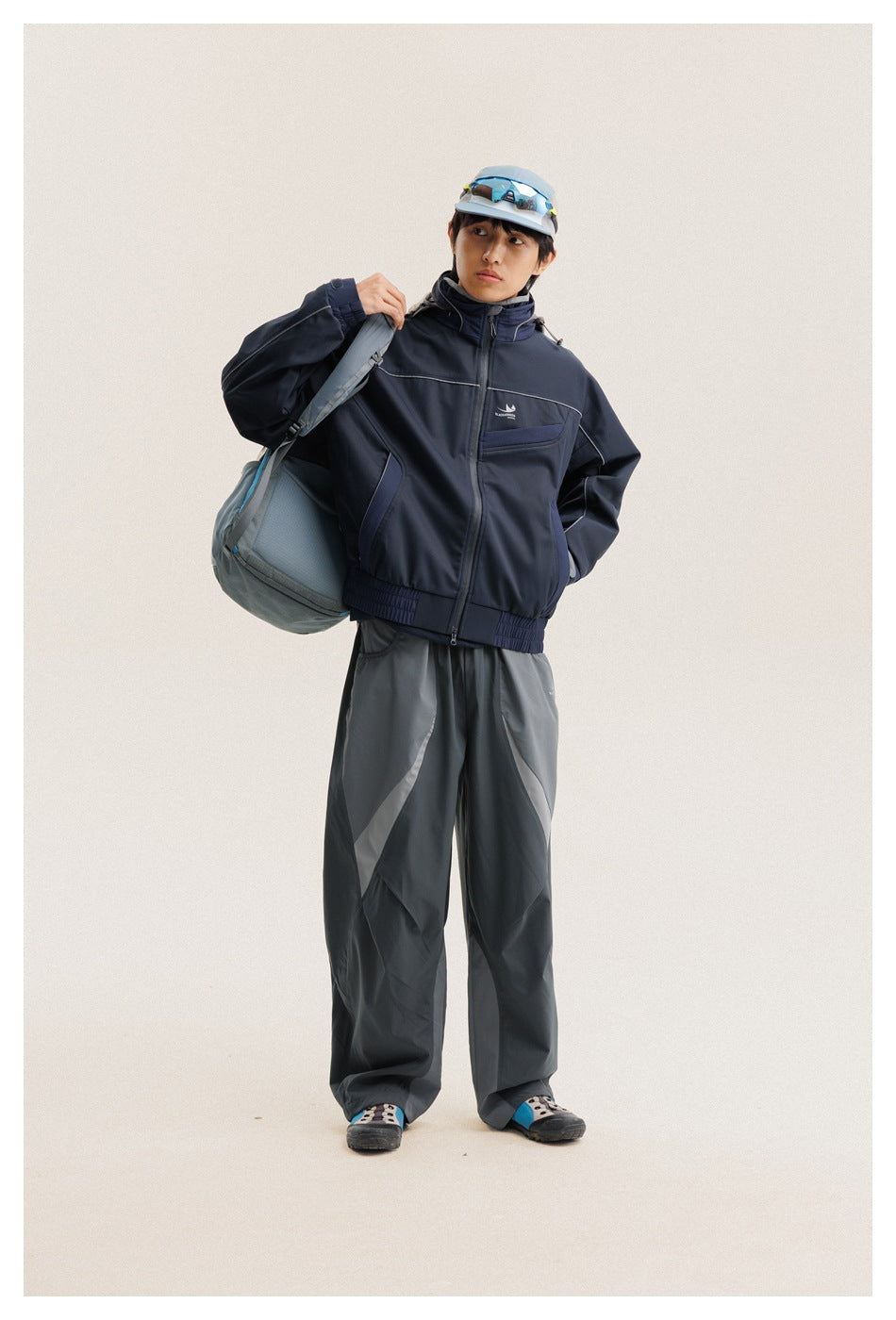 Full-length front view of a model wearing a navy hooded jacket and loose gray paneled trousers with a shoulder bag.