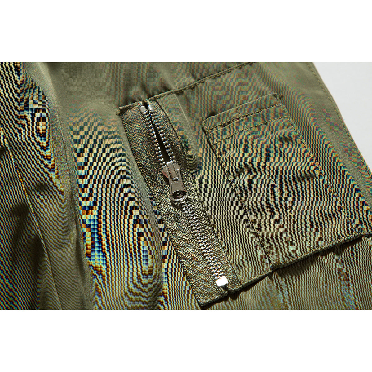 Close-up of an olive jacket sleeve pocket with a silver zipper and stitched flap.