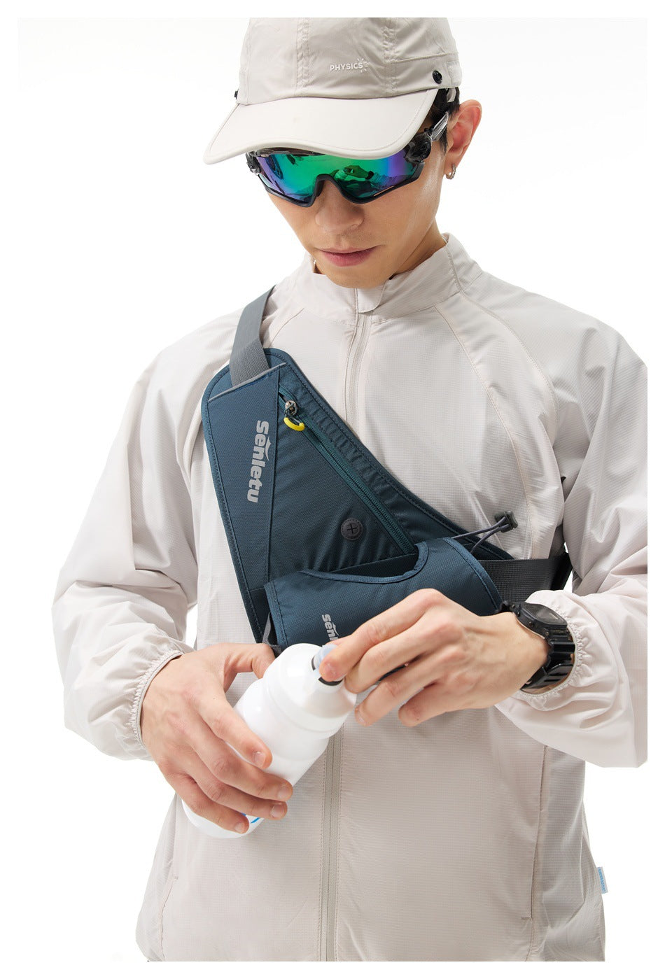 Close view of a model in a light gray zip jacket, wearing a blue chest bag and holding a white bottle.