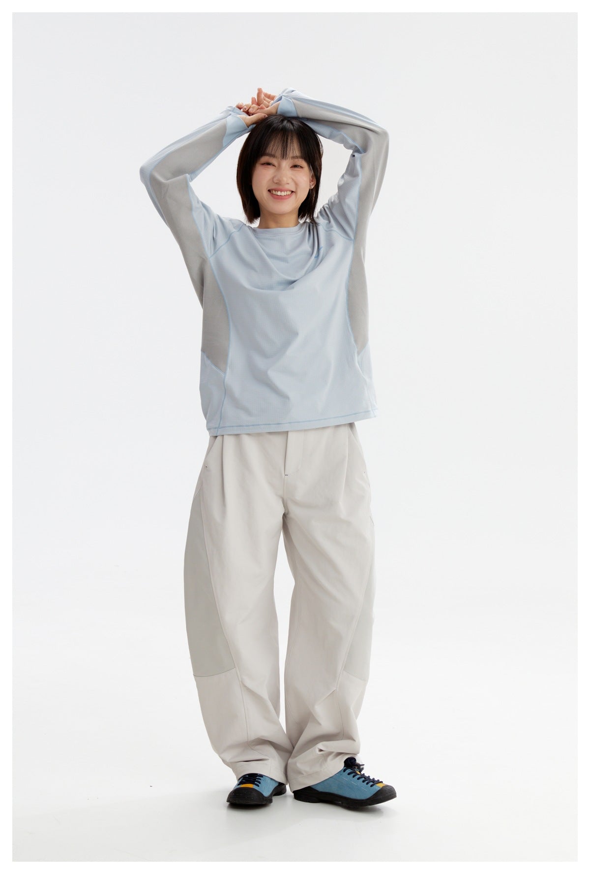 Model wearing a pale blue and gray long-sleeve shirt with white trousers, posing with arms raised.