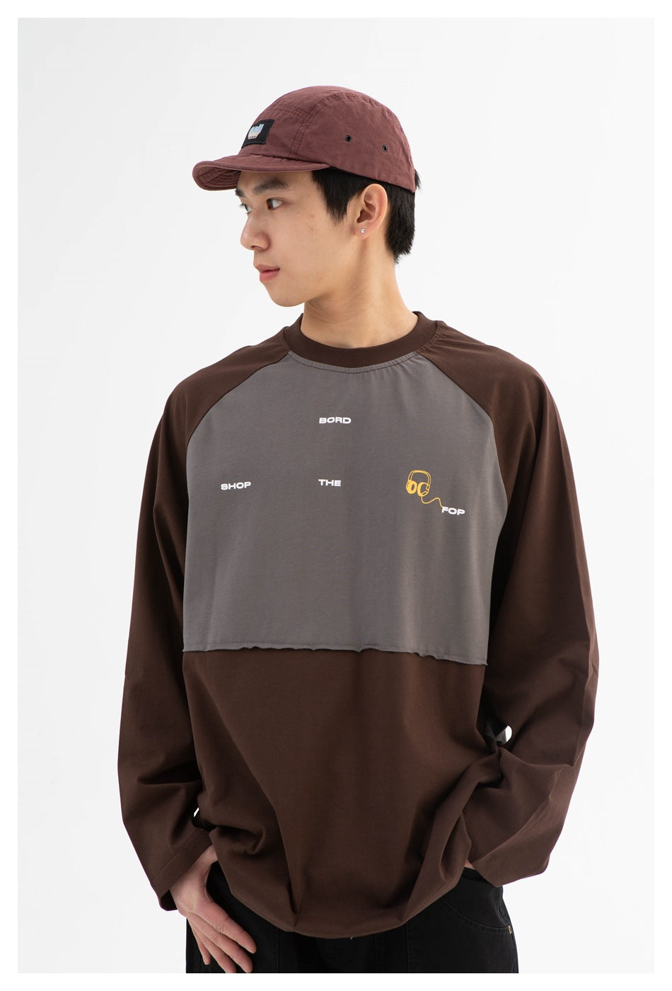 Close view of a model wearing a brown and gray color-block shirt with small chest graphics and a burgundy cap.