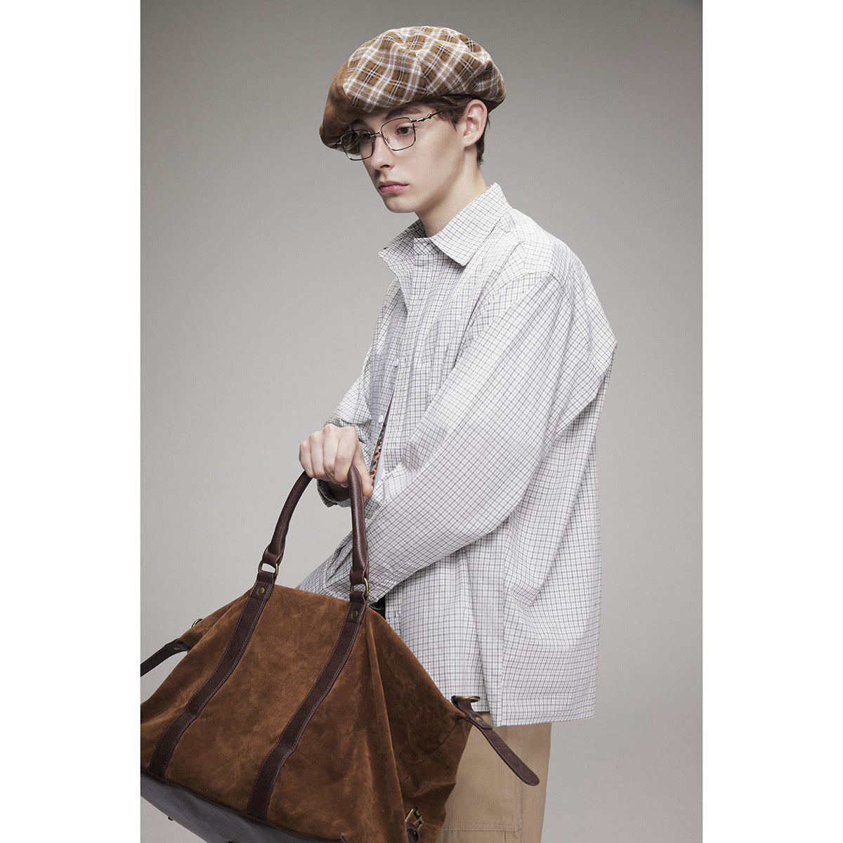 Close view of a model wearing an oversized light gray checked shirt, with a brown plaid cap and large brown bag.