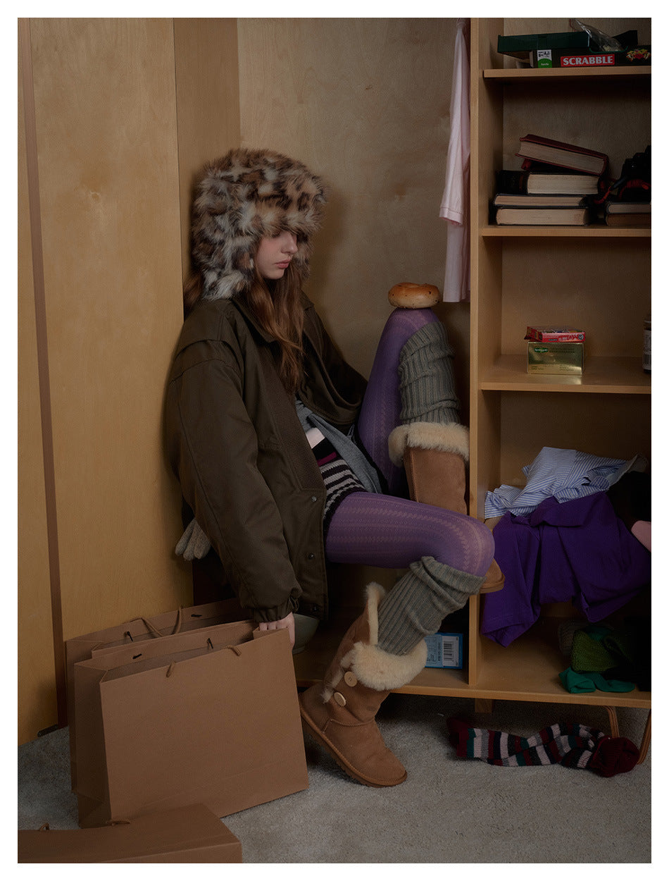 Seated model wearing an oversized olive-brown jacket and a spotted faux-fur trapper hat.