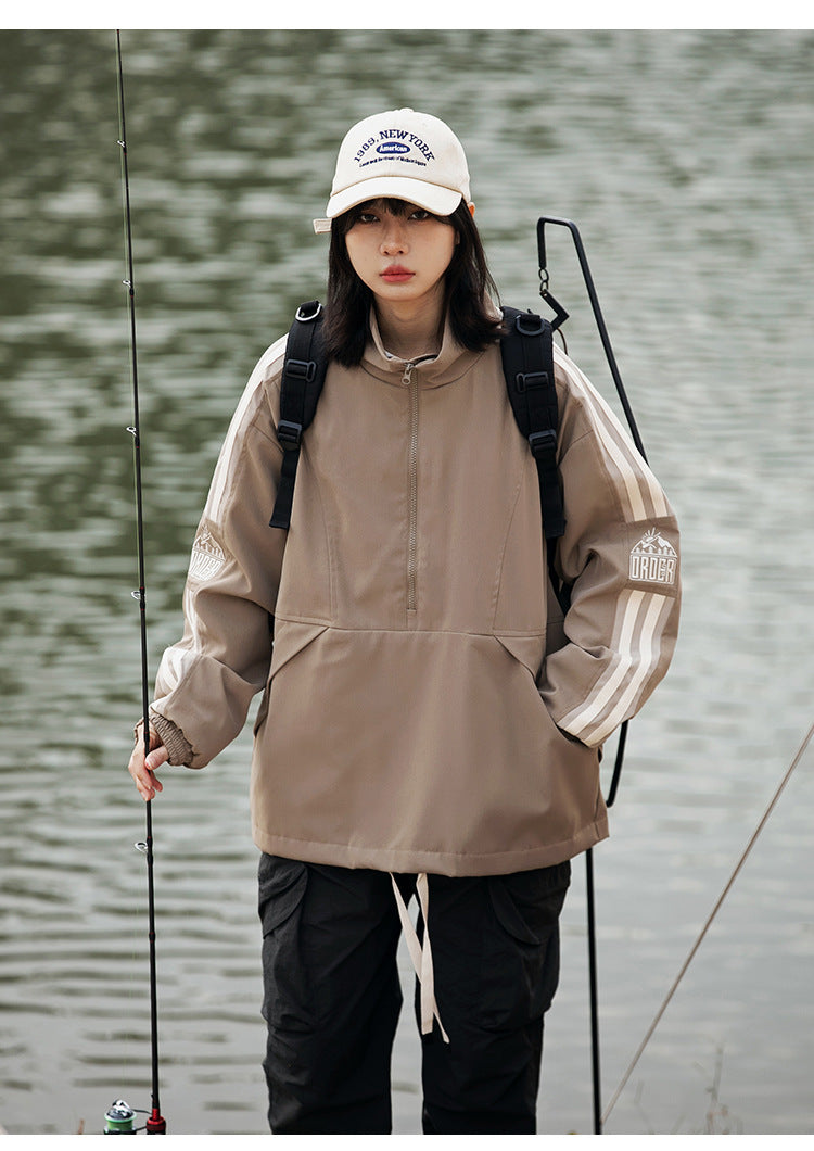 Model wearing a taupe pullover jacket with cream sleeve stripes and black pants beside a lake.