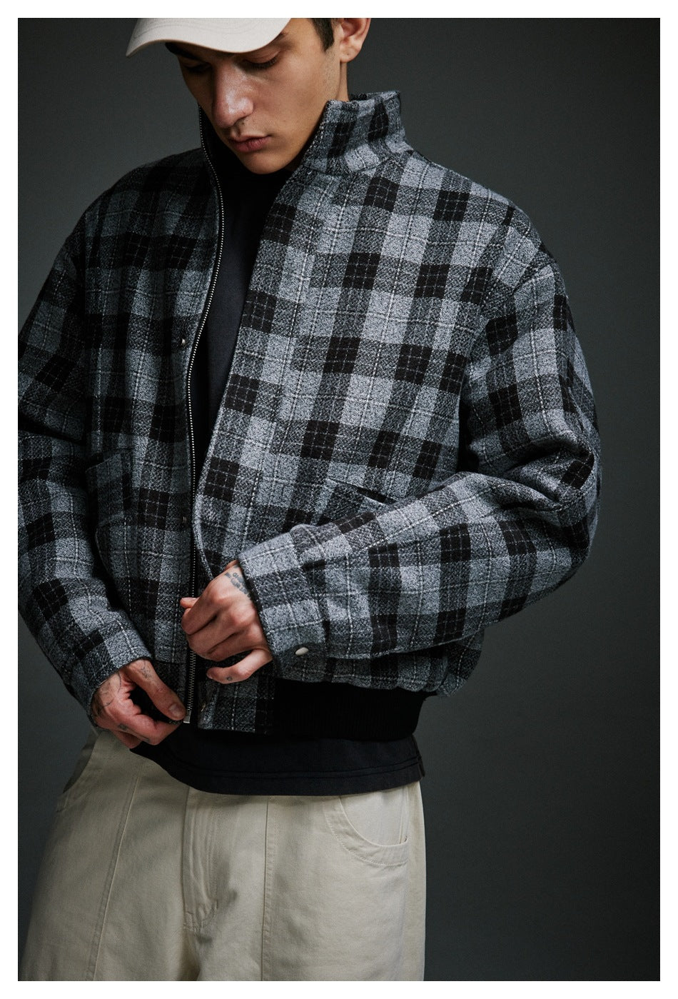 Close-up of a model adjusting a gray-and-black plaid zip jacket, with its collar, zipper, cuff, and ribbed hem visible.