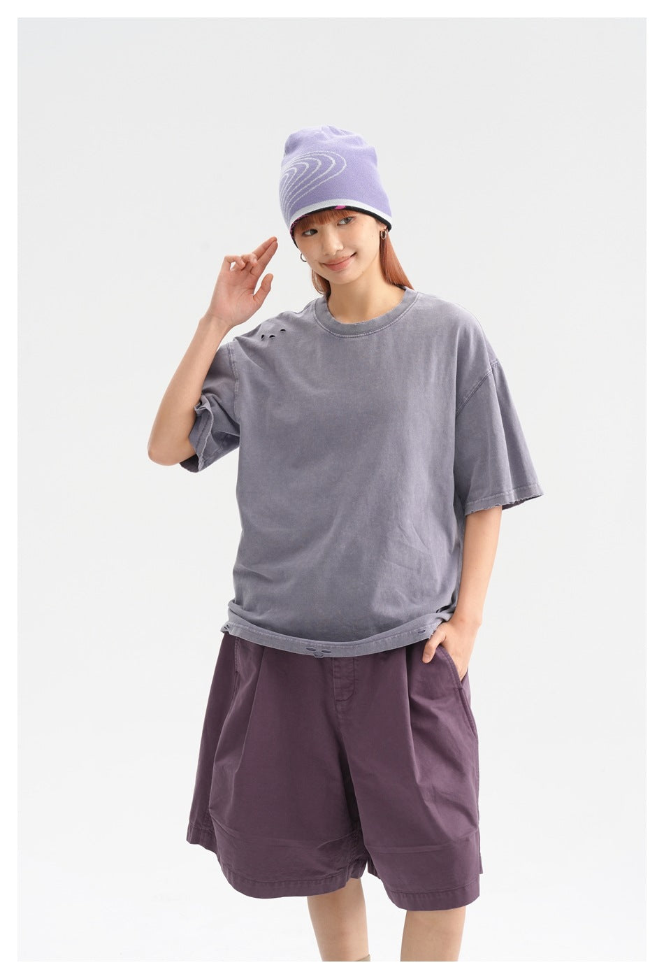 Model wearing an oversized faded lavender-gray T-shirt with small shoulder distressing and plum shorts, shown from the front.