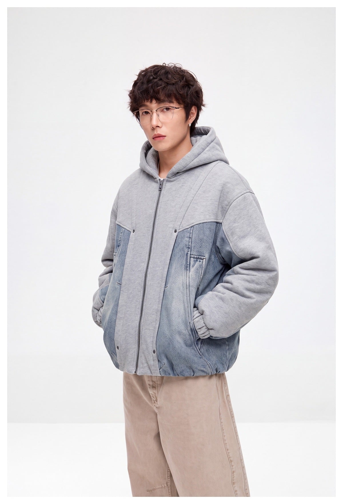 Angled front view of a model wearing a gray hooded zip jacket with blue denim panels and beige trousers.