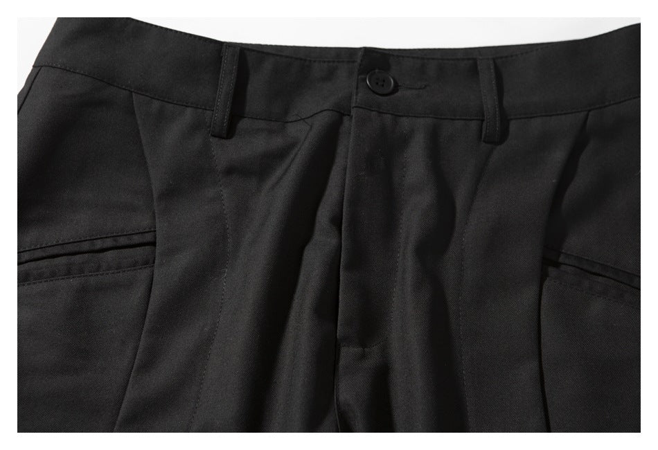 Close-up of black trousers showing the buttoned waistband, belt loops, fly, pleats, and pocket openings.