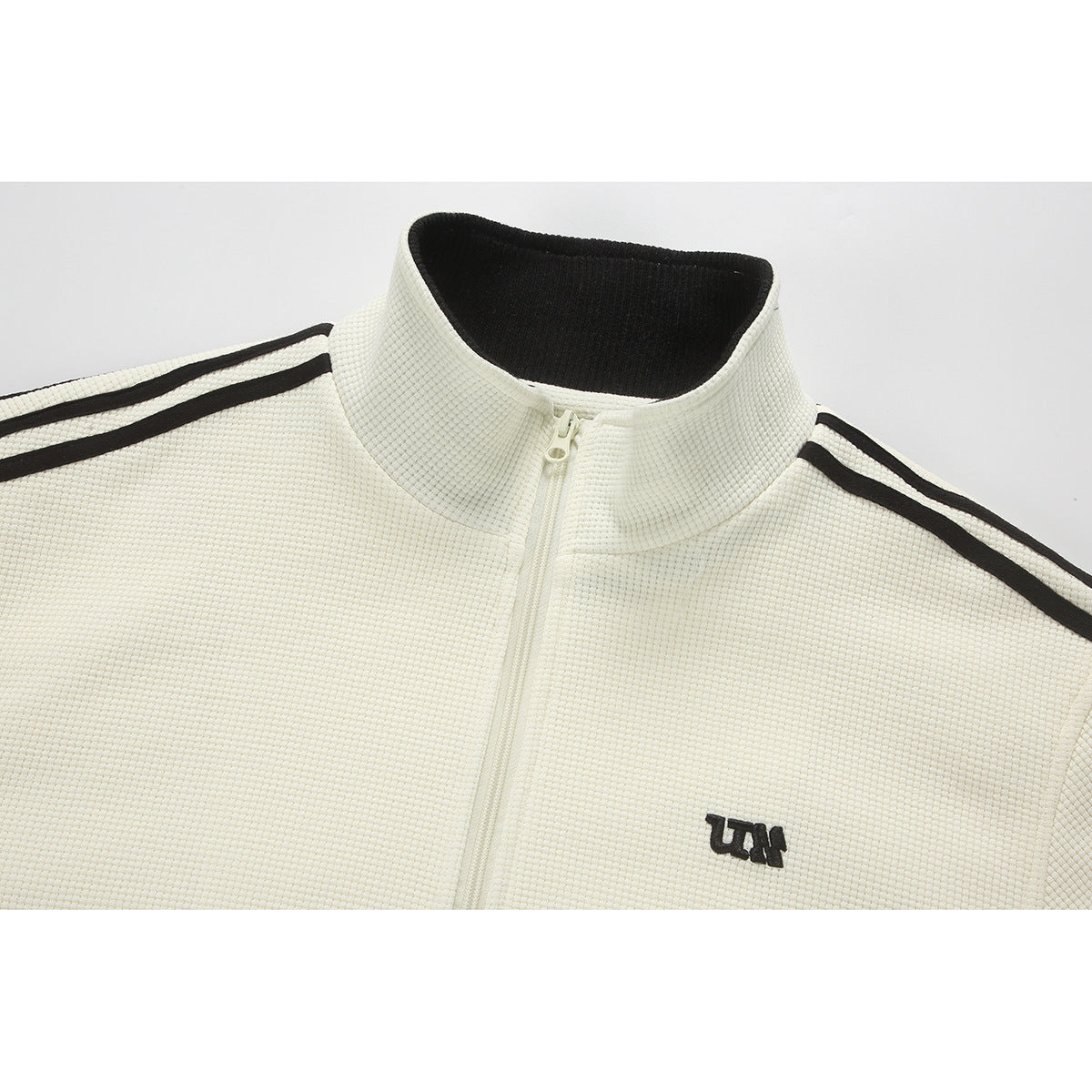 Close-up of an ivory zip-up jacket with a black inner collar, black shoulder stripes, and a small embroidered chest logo.