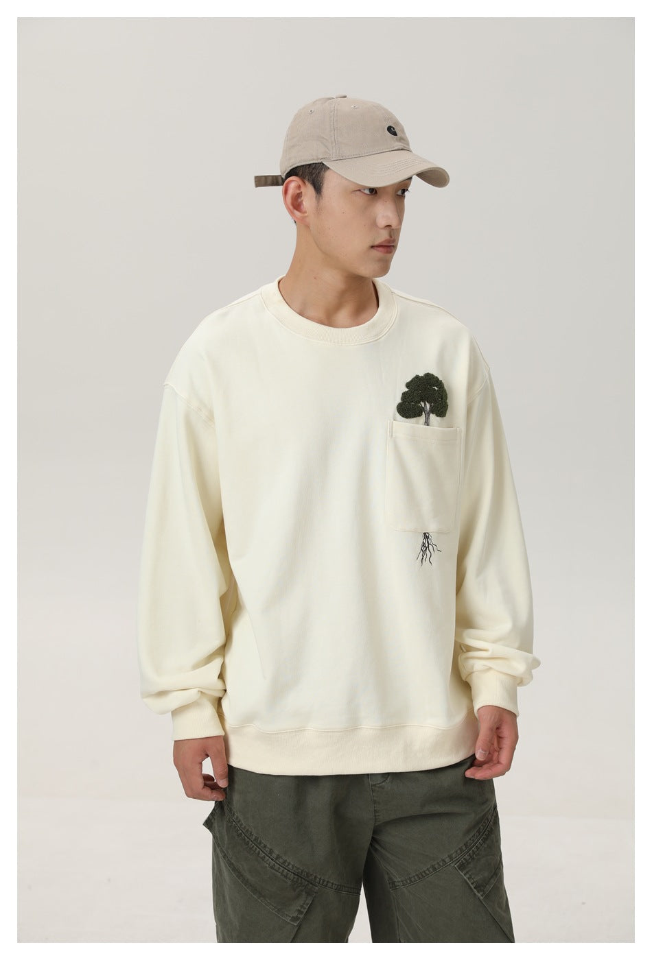 Model wearing an ivory crewneck sweatshirt with a chest pocket and embroidered tree-and-roots design.