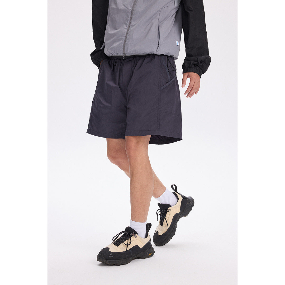 Model wearing dark charcoal drawstring shorts with angled pockets and a gray-and-black jacket.