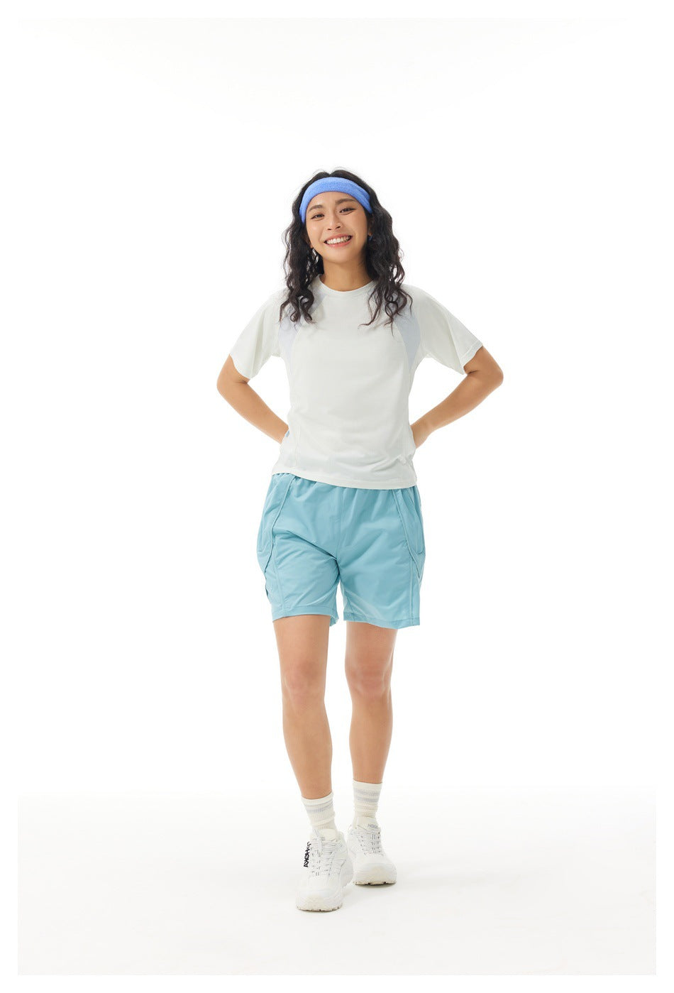 Woman in a white raglan T-shirt with gray-blue panels, blue shorts and headband, standing with hands on hips