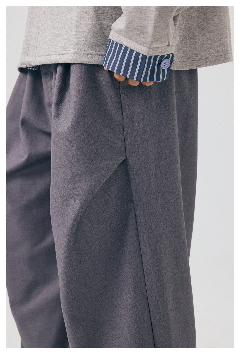 Close-up of loose gray trousers beneath a light gray top, with a blue striped buttoned cuff.