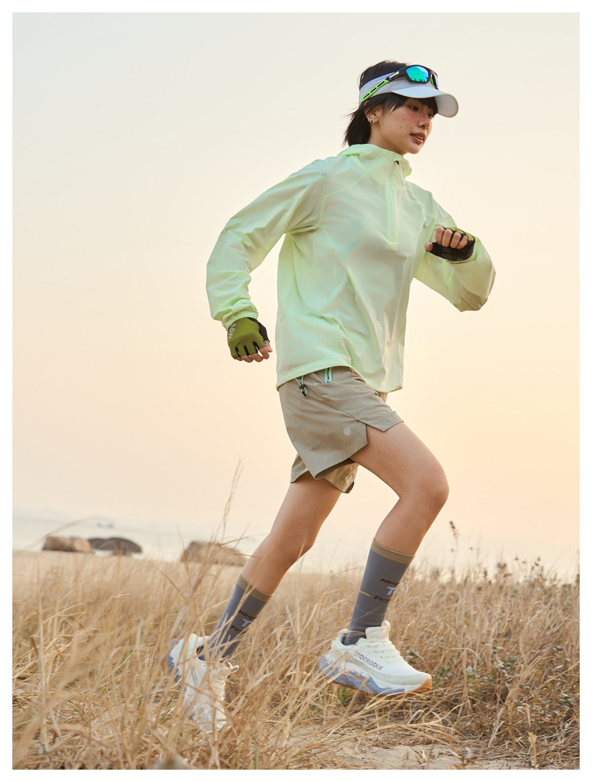 Runner in a pale lime quarter-zip pullover and taupe shorts moving through coastal grass at sunset.