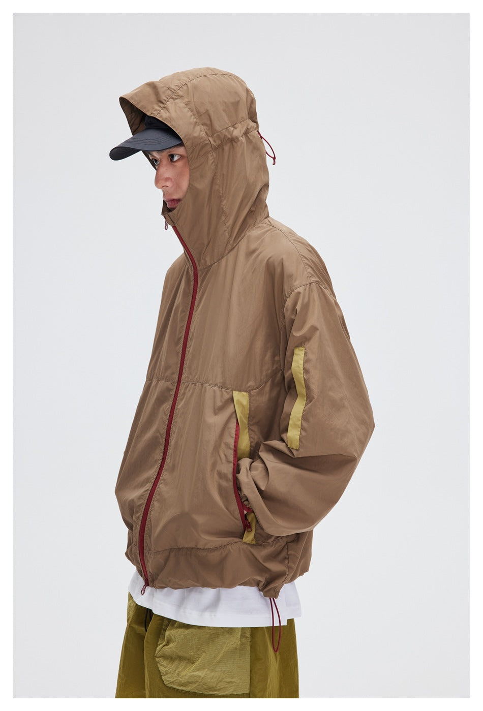 Side view of a model wearing a taupe hooded jacket with a red zipper and yellow accents.