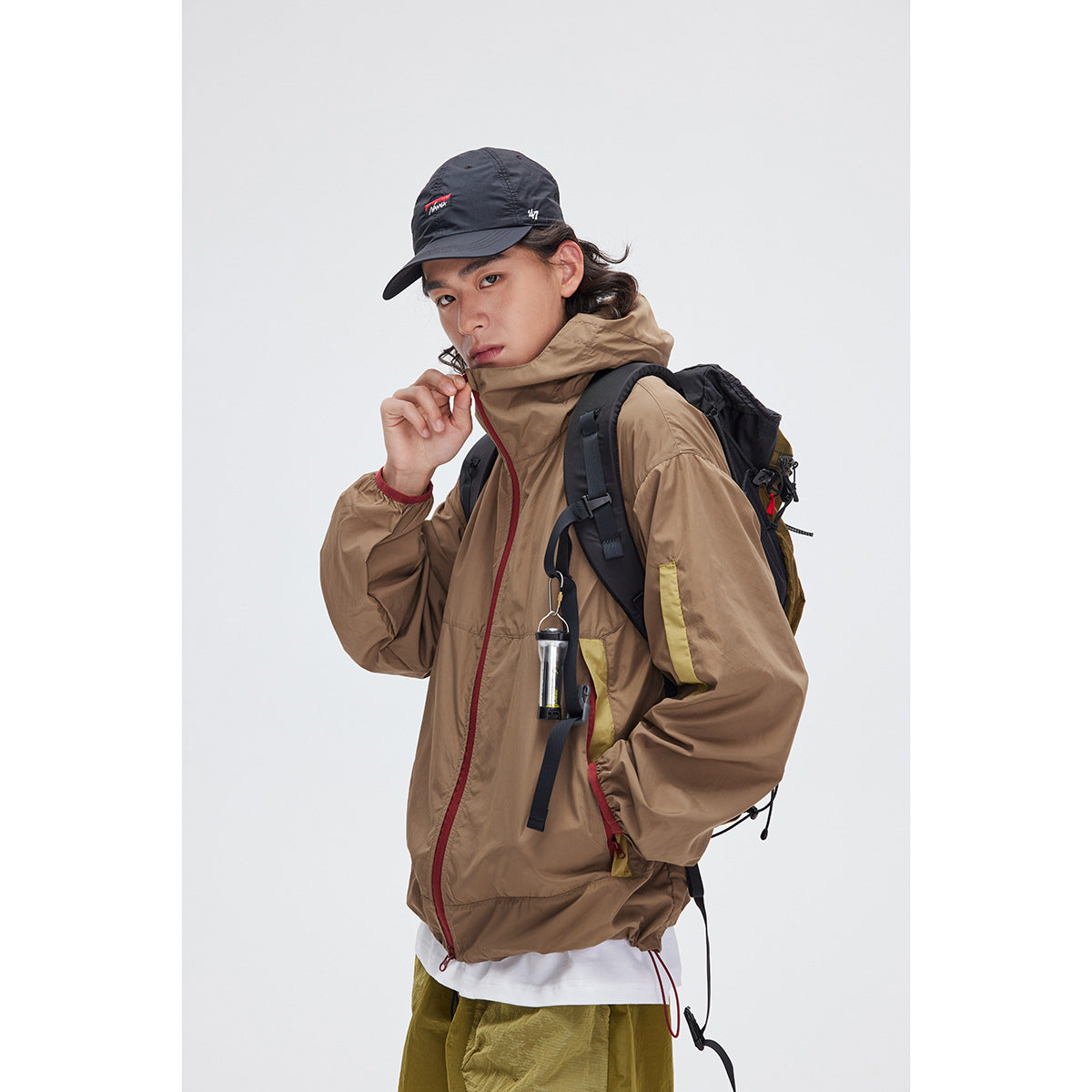 Model wearing a taupe hooded jacket with red zipper trim, shown from the front with a backpack.