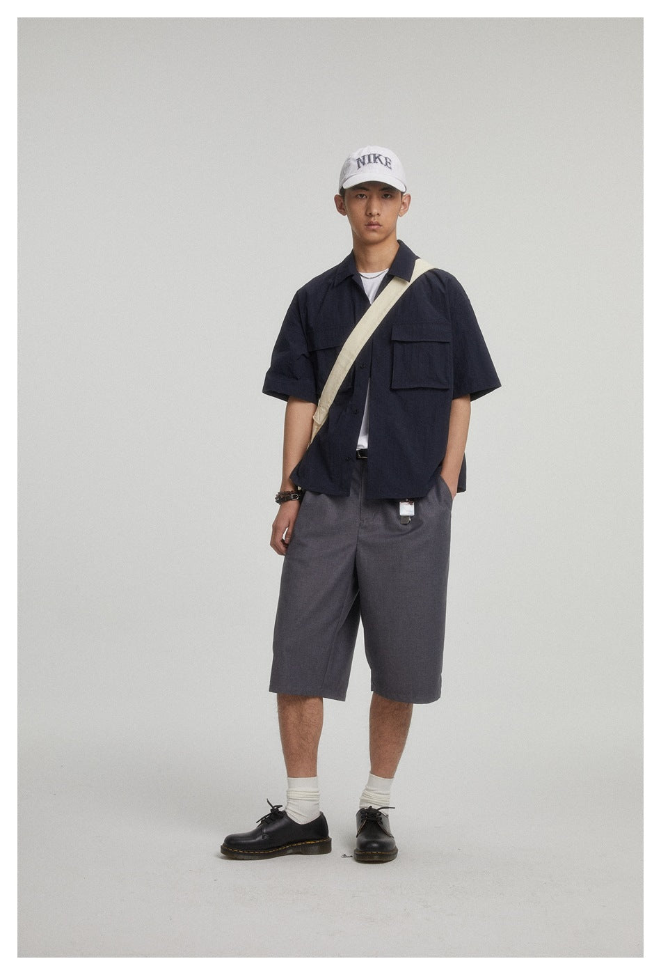 Model wearing gray pleated knee-length shorts with a navy short-sleeve shirt, white cap and black shoes