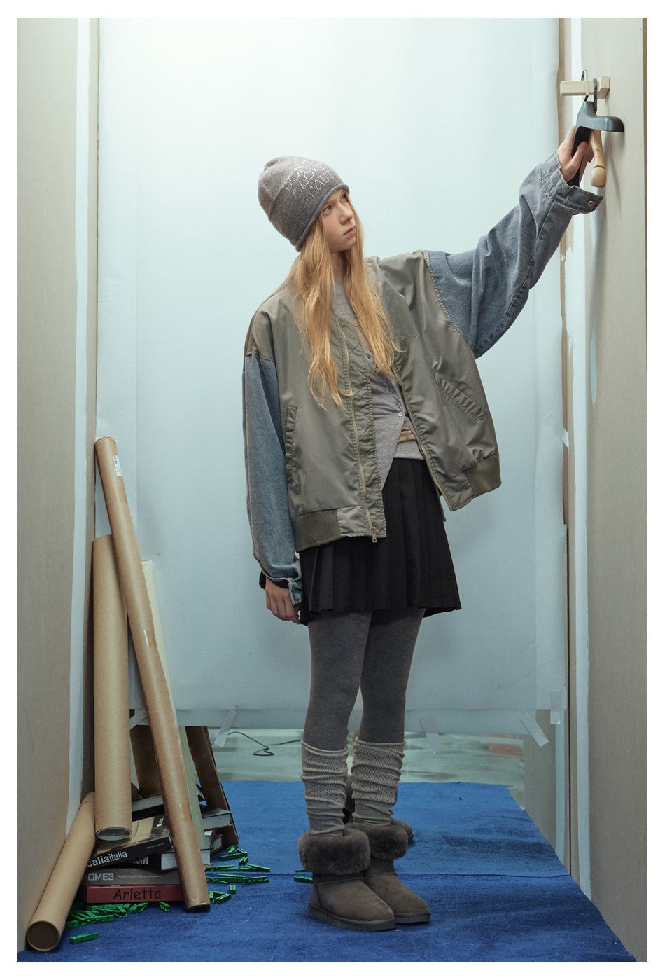 Model reaching up to a wall hook in an olive-gray bomber jacket with denim sleeves, black pleated skirt and boots