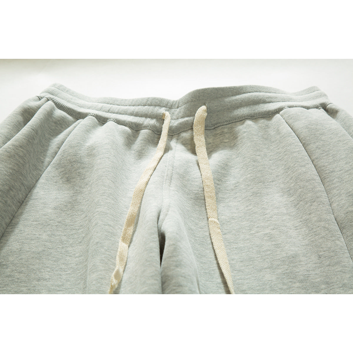 Close-up of light gray sweatpants showing the elastic waistband, cream drawstrings, and front seams.