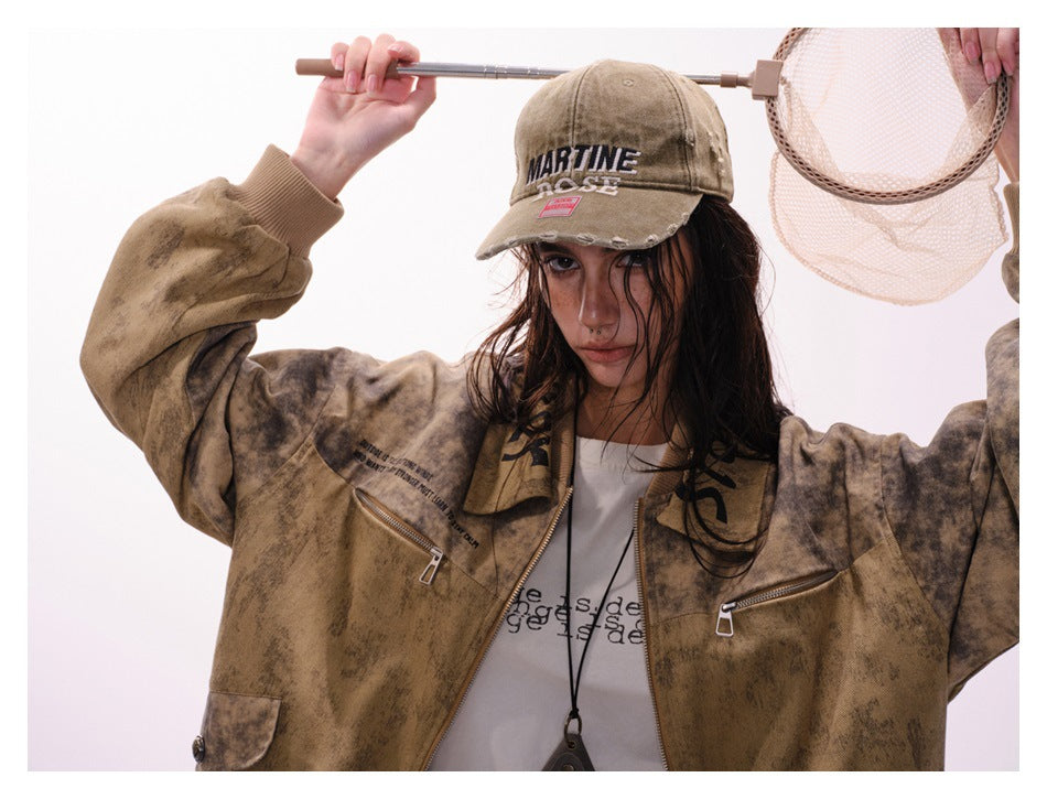 Model wears a mottled brown zip-front jacket with chest zippers and a distressed tan cap, holding a mesh net.