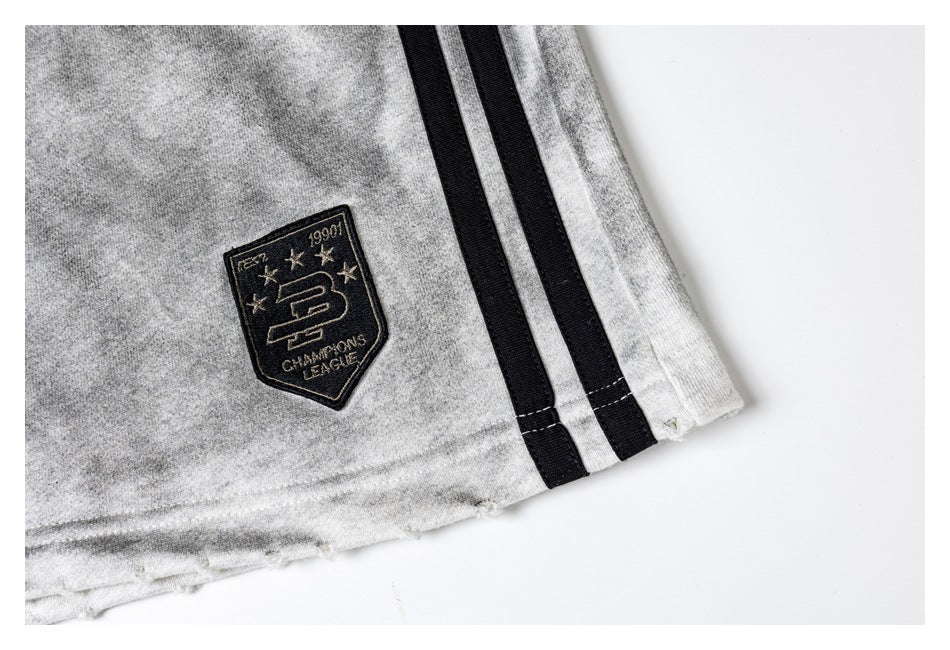 Close-up of light gray shorts with black side stripes and a black embroidered shield patch.