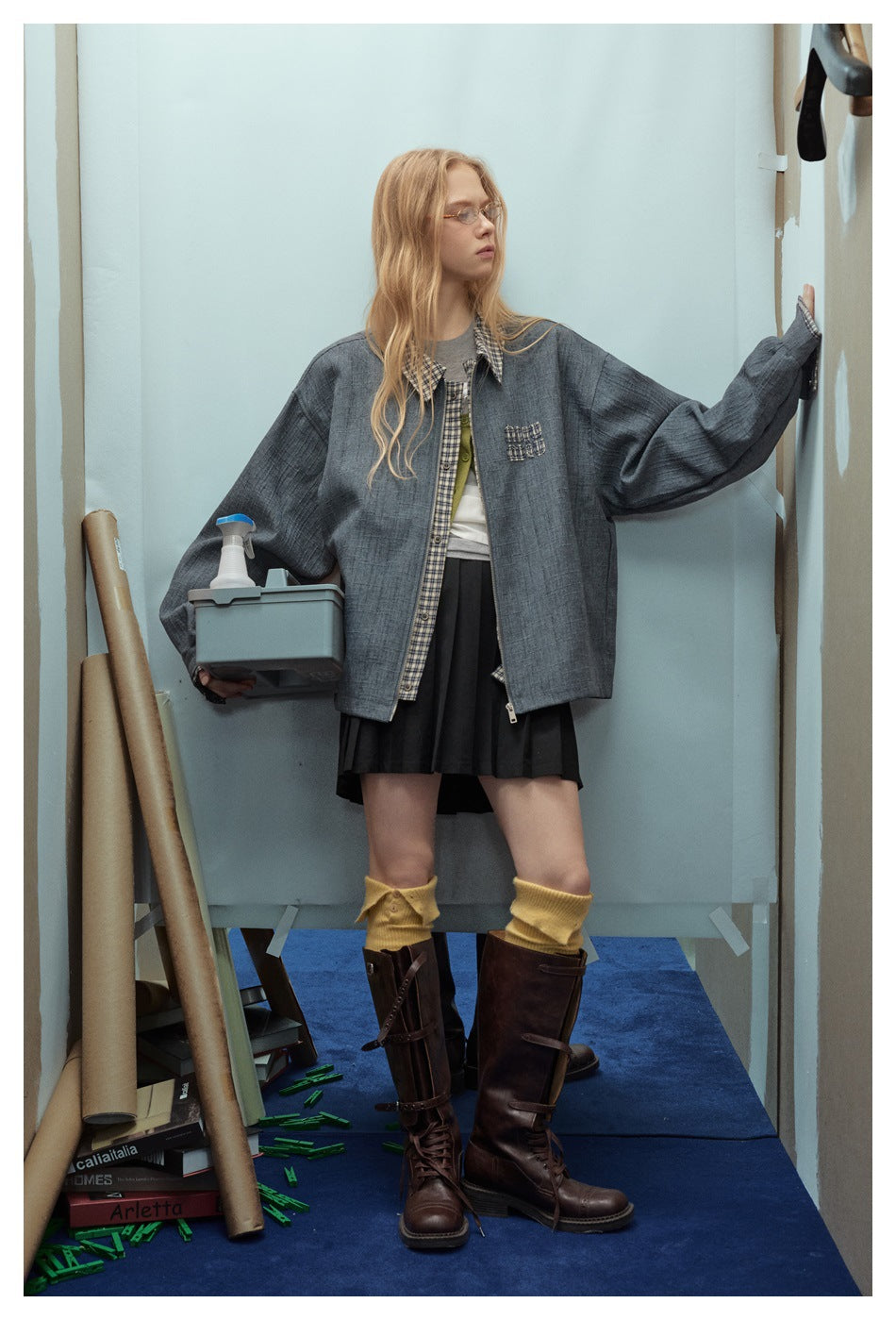 Model wearing an oversized blue-gray jacket with plaid trim and chest pocket, black skirt, yellow socks, and brown boots.