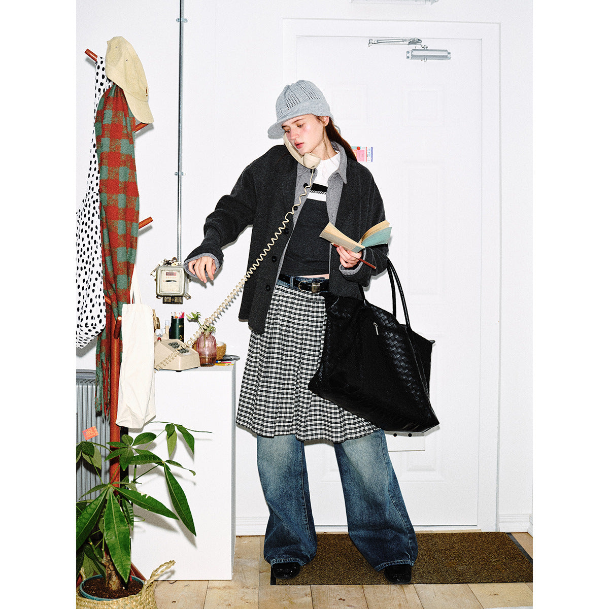Model wears a charcoal button-front jacket, gray knit hat, gingham skirt layered over wide-leg jeans, and carries a black